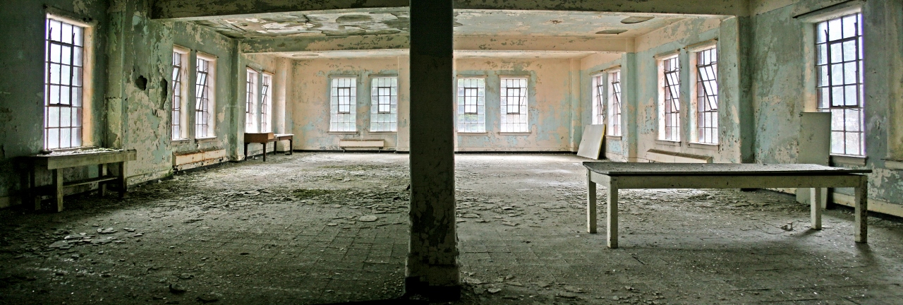 A room with crumbling, peeling walls. Table on the right, a bench on the left. Another smaller table can be seen at the back of the room