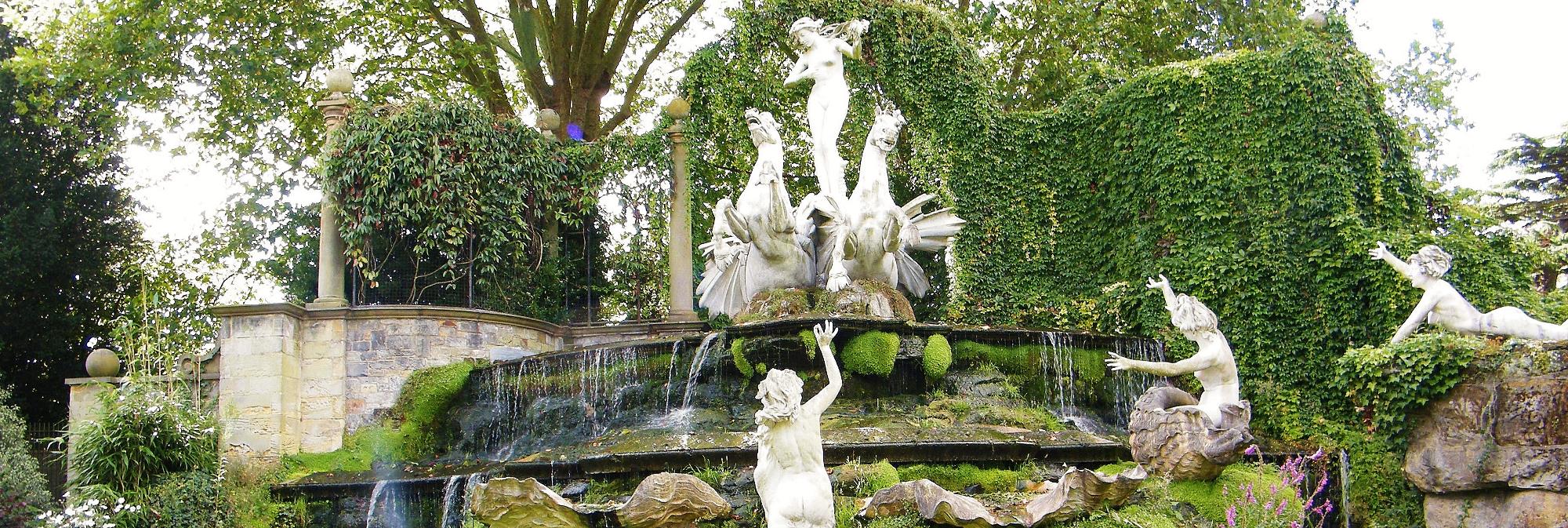 Big fountain with two white horse statues. Three women statues around leaning towards the fountain. Surrounding looks like in the park