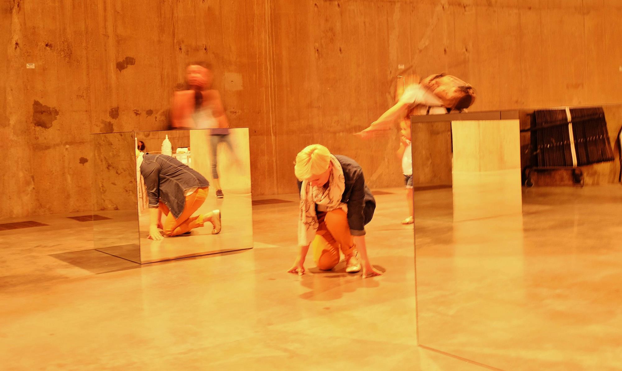 Woman kneel down on the ground, looks like a performance, mirror next to her. There are more people around, but they are blurred